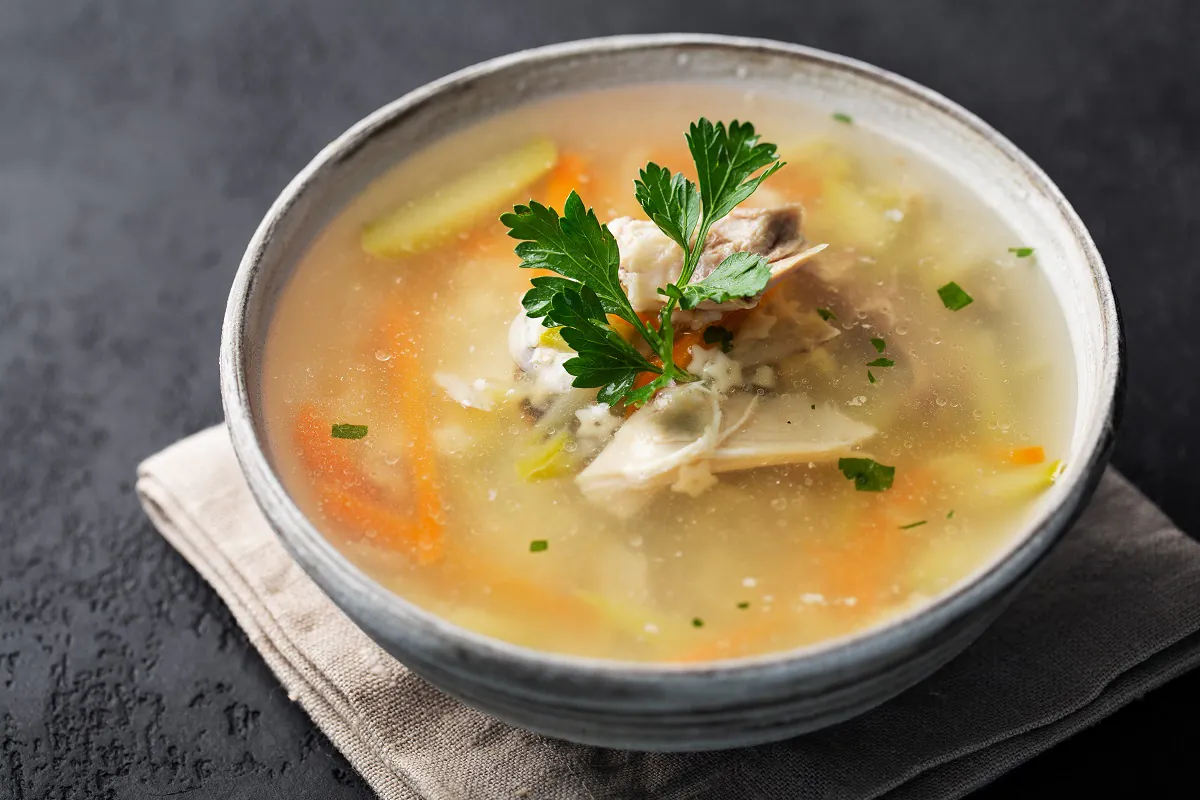 Steaming bowl of cock soup on a rustic table with fresh herbs and vegetables.